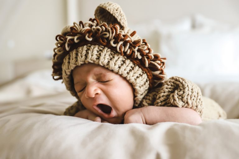 Newborn baby yawning while wearing a cozy knitted lion outfit, lying on a soft bed