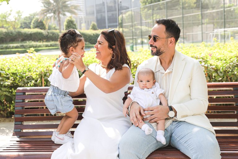 Happy family enjoying a sunny day in a park in Dubai, with a mother playing with her daughter, father holding a baby, all dressed in white